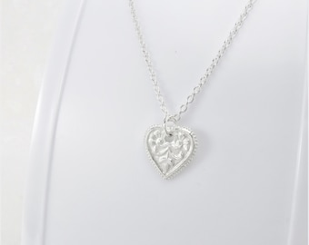May include: A silver necklace with a delicate, ornate heart pendant. The heart is detailed with floral designs and a beaded border.