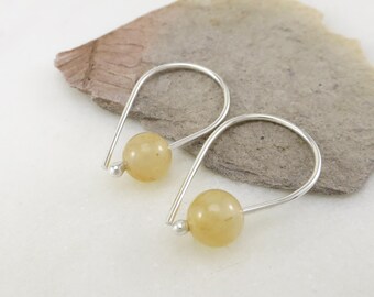 May include: A pair of silver-toned hoop earrings with a small, pale yellow bead at the bottom and a larger, matching bead at the center. The earrings are displayed on a gray stone surface.