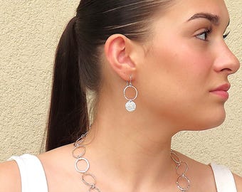 May include: Silver earrings with a hammered circle pendant and a hoop. The earrings are hanging from the earlobe of a person with long dark hair.