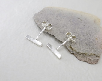 May include: A pair of silver bar stud earrings. Each earring features a rectangular bar attached to a post with a butterfly backing. The earrings are displayed on a gray, textured surface.