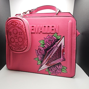 May include: Pink leather handbag with a  "EVADDEN" logo and a painted design of a pyramid with roses. A small pink pouch with a red circle design is attached to the bag.