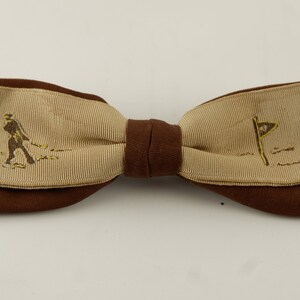 Può includere: Papillon marrone con una sezione centrale beige con un'immagine di un golfista e una bandiera da golf. Il papillon è realizzato in tessuto a coste e ha un nodo centrale e bordi marrone scuro. Un accessorio elegante per gli appassionati di golf.