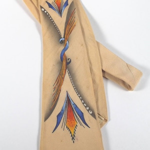 May include: A tan necktie featuring a hand-painted design with two symmetrical blue and orange geometric floral motifs connected by a central blue swirl and a curved line of white dots, creating a decorative pattern on the fabric surface.