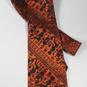 May include: A necktie featuring a vibrant orange background with a repeating pattern of black abstract symbols and geometric shapes. The design includes stylized figures, circular dots, and linear borders that run vertically along the fabric.