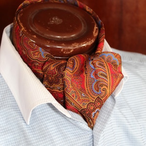Puede incluir: Un pañuelo ascot de color rojo, marrón y azul con estampado de cachemira está anudado y se lleva con una camisa azul claro y un cuello blanco. Un objeto redondo marrón está detrás del pañuelo. El pañuelo tiene un patrón ornamentado.