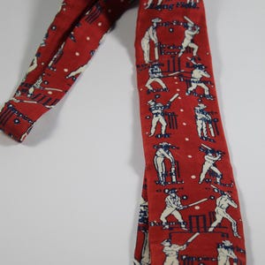 May include: Red necktie with a repeating pattern of cricket players in white and navy blue. The tie features a classic design with a pointed end and a repeating pattern of cricket players in action.