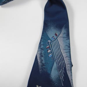 May include: A navy blue tie with a geometric design in shades of teal and silver. The tie features a small, stylized bird emblem near the tip. The design includes vertical stripes and rectangular shapes.