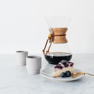 Op de afbeelding: Een glazen koffiezetapparaat met een houten kraag en leren riem, gevuld met donkere koffie. Twee grijze keramische bekers en een stukje blueberry cheesecake op een bord met een gouden vork.