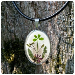 Könnte beinhalten: Eine silberne ovale Anhänger-Halskette mit einem schwarzen Lederband. Der Anhänger enthält ein gepresstes vierblättriges Kleeblatt und andere kleine grüne und violette Blumen.