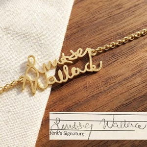 May include: A gold-colored necklace with the name "Lussey Wallace" in a cursive font. The necklace is on a wooden surface with a white cloth. A small paper with the signature "Lussey Wallace" is also present.