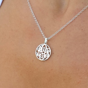 以下が含まれることがあります： 複雑なモノグラムデザインが施された円形ペンダントが付いたシルバーネックレス。ペンダントは繊細なチェーンから吊り下げられています。白いトップスを着ている人。