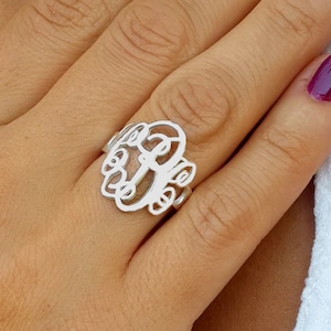 Könnte beinhalten: Ein silberner Monogrammring mit einem kunstvollen, durchbrochenen Design. Der Ring zeigt drei stilisierte Initialen. Der Ring befindet sich an einem Finger mit lackierten Nägeln. Weißer Hintergrund.