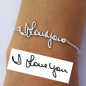 May include: A silver bracelet with the words 'I love you' written in cursive. The bracelet is on a person's wrist. A white piece of paper with the same words written in cursive is below the bracelet.
