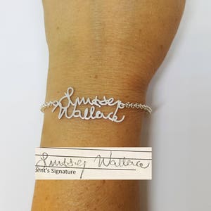 May include: A silver-toned metal bracelet featuring a custom cursive script design that reads Lindsey Wallace. The bracelet is attached to a delicate chain and is displayed alongside a white paper strip showing the original signature Lindsey Wallace.