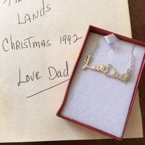 May include: A gold-toned metal necklace featuring a cursive script pendant that reads Love Dad, resting inside a small red gift box with a white interior lining. The pendant is attached to a delicate silver-toned chain on both sides.