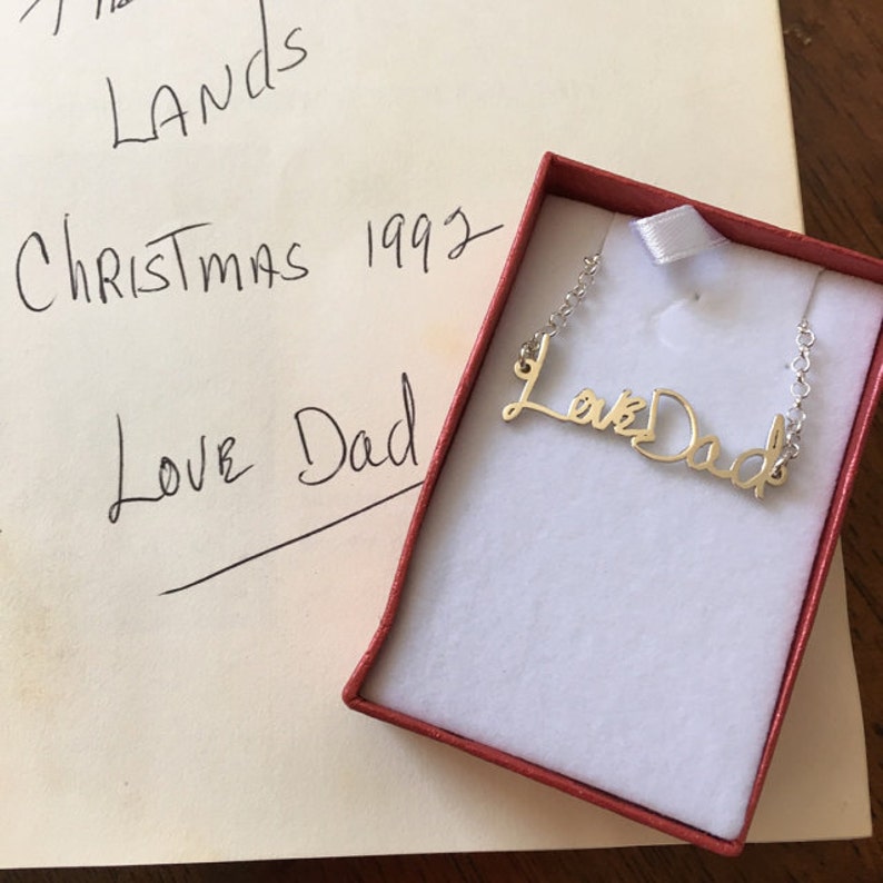 May include: A gold-toned metal necklace featuring a cursive script pendant that reads Love Dad, resting inside a small red gift box with a white interior lining. The pendant is attached to a delicate silver-toned chain on both sides.