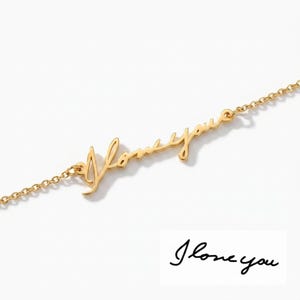May include: A delicate gold-colored necklace with the cursive phrase "I love you" as the centerpiece. The chain is fine and links to the scripted words. A handwritten "I love you" is also shown below the necklace.