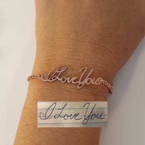 May include: A rose gold-toned bracelet featuring a cursive script metal cutout that reads "Love You". Below the bracelet, a piece of white lined paper displays the handwritten text "I Love You" in blue ink, matching the script style of the bracelet.