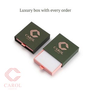 May include: Two square jewelry boxes with dark green lids and light pink sliding trays. The lids feature a rose gold logo with the text CAROL JEWELRY ART. The text Luxury box with every order is displayed above the boxes, and the logo is below.