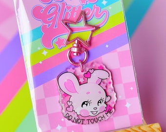 May include: A clear acrylic keychain featuring a pink cartoon rabbit with large eyes and heart accents, attached to a star-shaped metal clasp. The backing card displays the text GUTS & GLITTER and the phrase DO NOT TOUCH ME on the rabbit charm.