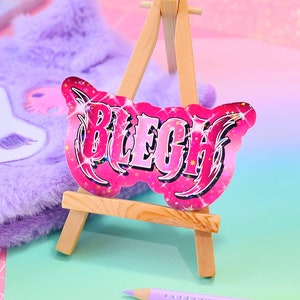 以下が含まれることがあります： スタイリッシュなフォントで「BLEGH」と書かれたピンクと白のキラキラしたステッカー。ステッカーは小さな木製イーゼルの上に置かれています。