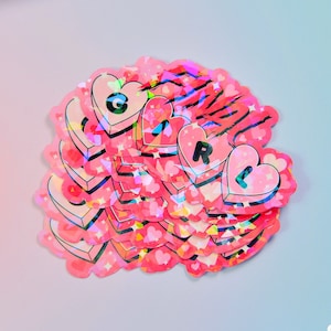 Può includere: Un mucchio di adesivi olografici rosa con la parola "GIRL" scritta in lettere rosa. Ogni lettera è su un adesivo separato a forma di cuore con uno sfondo scintillante. Gli adesivi sono disposti in una pila con le lettere visibili. Gli adesivi sono fatti a mano e hanno un logo "Guts & Glitter" nell'angolo in basso a destra.