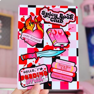 May include: A pink and black chequered background with stickers featuring a heart with flames, a book with flames, a knife, and a stack of books with the text "Spicy Book Club", "Dark Romance", "Explicit Content", "Touch Her & Die", "Trigger Warning", and "Hello, I'm Reading Smut".