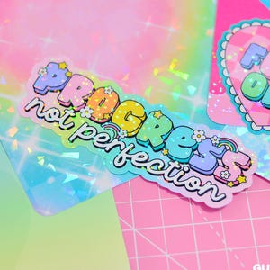 May include: A holographic sticker with the words "Progress not perfection" in pastel colours, decorated with flowers and rainbows. The sticker is on a colourful background with other stickers and a pink grid.