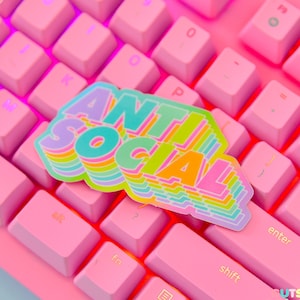 May include: A sticker with the words "Anti Social" in a rainbow gradient font. The sticker is placed on a pink keyboard.