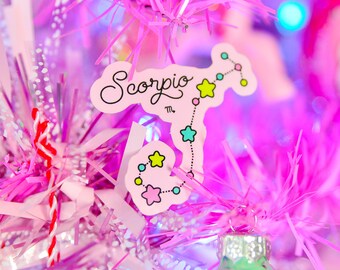 May include: A pink Christmas tree ornament with a white background and the word "Scorpio" written in cursive. The ornament also features a constellation of stars in blue, yellow, and pink.