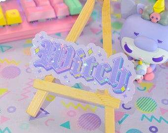 May include: A purple and pink glitter sticker with the word "Witch" in a gothic font. The sticker is on a small wooden easel and is sitting on a purple and pink patterned surface.