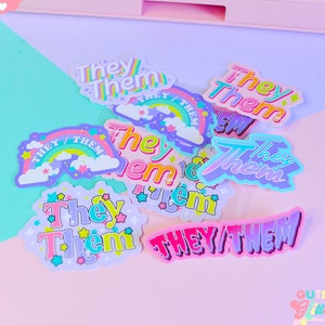 May include: A collection of colourful stickers with the phrase "They/Them" in various fonts and designs. Some stickers feature rainbows, stars, and clouds. The stickers are on a pink and light blue background.