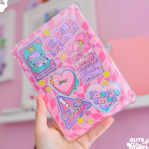May include: A pink clear phone case with various colourful stickers. The stickers include a heart with the word "SMUT" on it, a sign that says "Explicit Content", a sign that says "Please Do Not Disturb Reading In Progress", and a sign that says "Reading Hot Girl Shit".