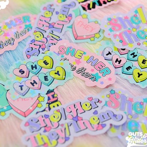 May include: A collection of colourful stickers featuring different pronoun combinations, such as "She/Her/They", "She/They", and "She/Her/Them". The stickers have heart, star, and glitter details. The text "GUTS & GLITTER" is visible in the bottom right corner.