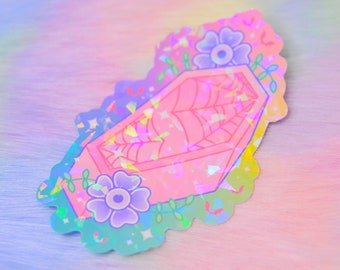 May include: A holographic sticker featuring a pink coffin with a spiderweb design, surrounded by purple flowers and green vines. The sticker has a scalloped edge and the words "Hand Made" and "Cuts & Glitter" are visible.