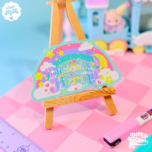 May include: A rainbow-colored sticker with glitter and stars, featuring the text "Basically a Hermit" in a colorful, retro font. The sticker is displayed on a small wooden easel.
