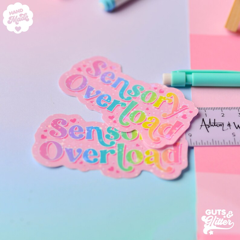 Pu&ograve; includere: Due adesivi rosa con testo glitter arcobaleno che recita "Sensory Overload". Gli adesivi sono su uno sfondo blu chiaro e rosa.