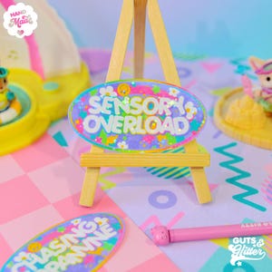 May include: A colorful oval resin sticker with the text "Sensory Overload" in white letters. The sticker is decorated with glitter, stars, and flowers. It is displayed on a miniature easel.