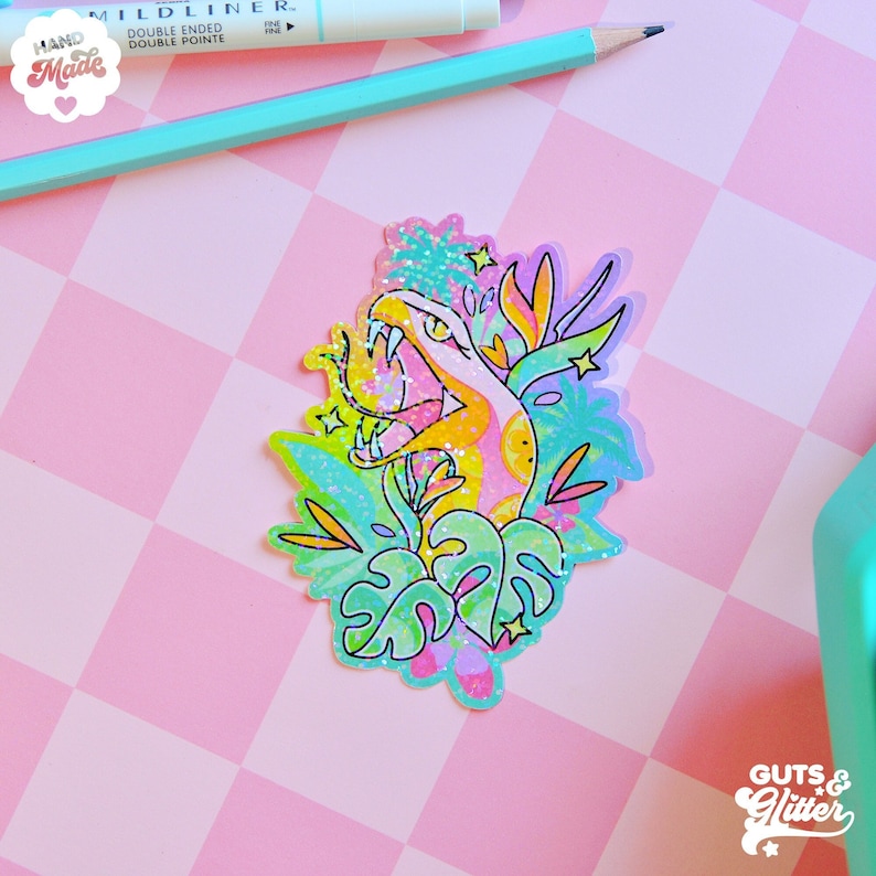 Pu&ograve; includere: Un adesivo colorato con un serpente di cartone animato con un corpo rosa, giallo e arancione, circondato da foglie tropicali verdi e blu e stelle. L'adesivo ha una finitura glitterata e il testo "GUTS & GLITTER" &egrave; visibile in basso a destra.