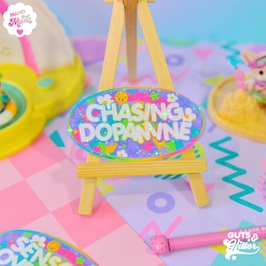 May include: A colorful, glittery, oval-shaped resin pin with the text "Chasing Dopamine" on a purple background. The pin is displayed on a small wooden easel.