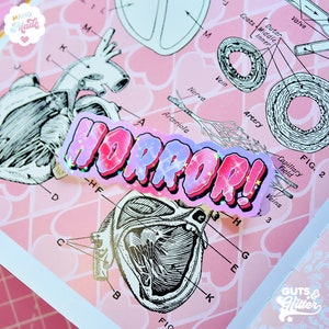 May include: A pink and purple glitter sticker with the word "HORROR!" in bold, white letters. The sticker is placed on a pink background with a vintage anatomical illustration of a heart.