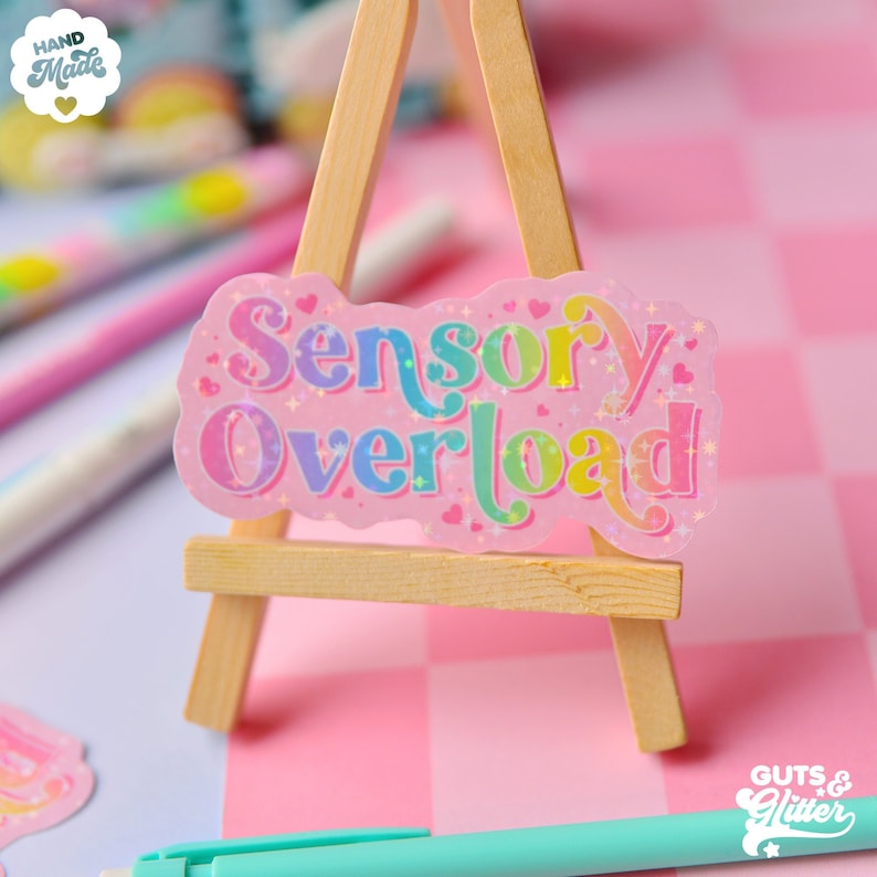 Pu&ograve; includere: Un adesivo rosa con glitter arcobaleno e il testo "Sensory Overload" con cuori e stelle. L'adesivo &egrave; su un piccolo cavalletto di legno.