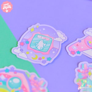 Op de afbeelding: Een paarse en roze sticker met een geest in een digitaal huisdier-achtig apparaat. De sticker heeft een witte wolk, een halve maan en sterren. De tekst "HAND Made" staat in de linkerbovenhoek.