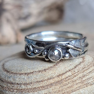 May include: A silver ring with a delicate, organic design featuring a grey, faceted gemstone. The ring is set on a textured, light brown surface.