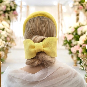 May include: A yellow headband with a large bow at the back. The headband is made of a woven material and is being worn by someone with blonde hair.