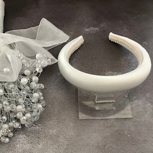 May include: A white satin headband with a padded interior. The headband is on a clear acrylic stand.