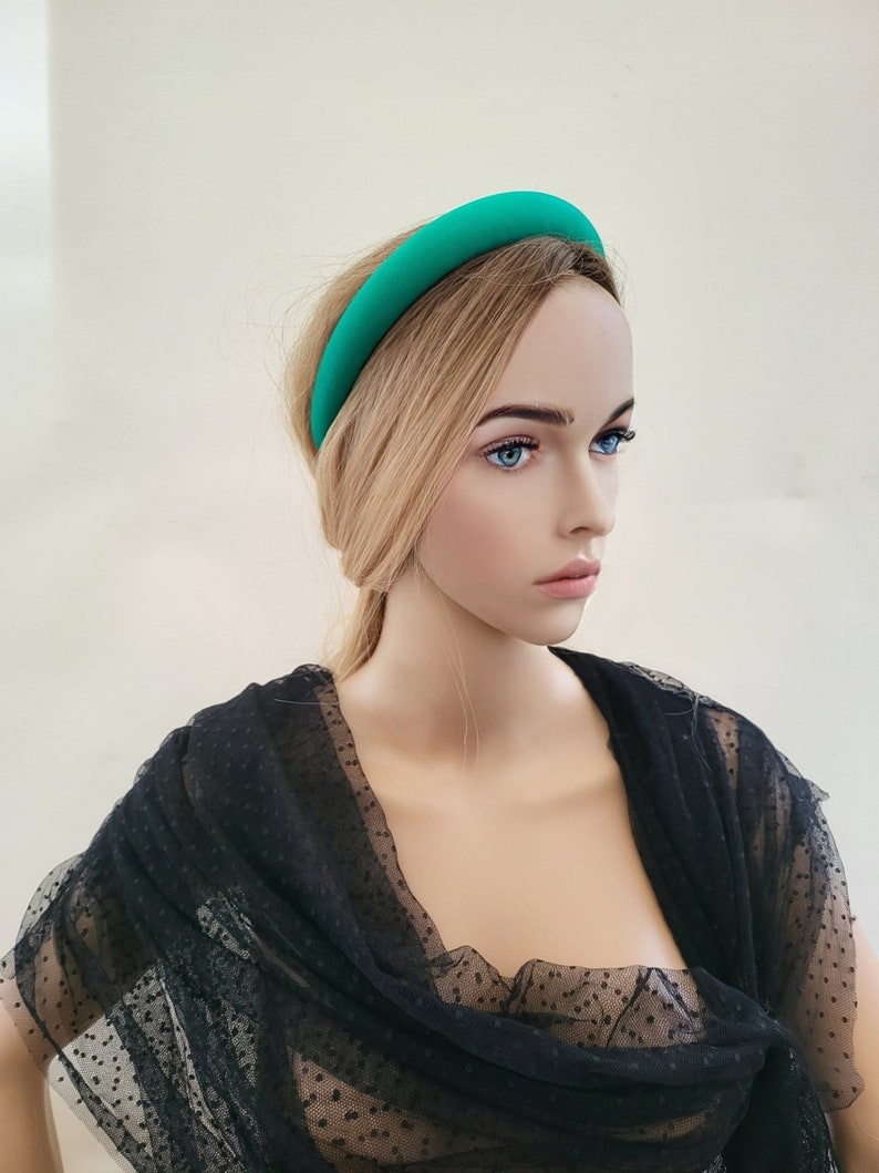 May include: A bright teal headband made of fabric. The headband is wide and has a smooth, rounded shape. It is worn by a mannequin with blonde hair and a black polka dot mesh top.