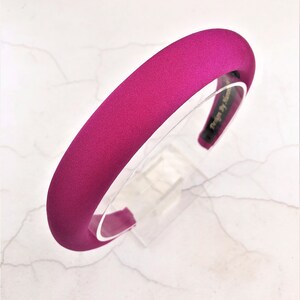 May include: A wide, fuchsia pink satin headband. The headband is made of a soft, smooth fabric and has a simple, elegant design.