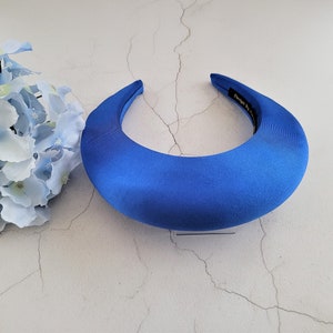 Può includere: Un cerchietto in raso blu brillante con una forma curva. Il cerchietto è su uno sfondo bianco.