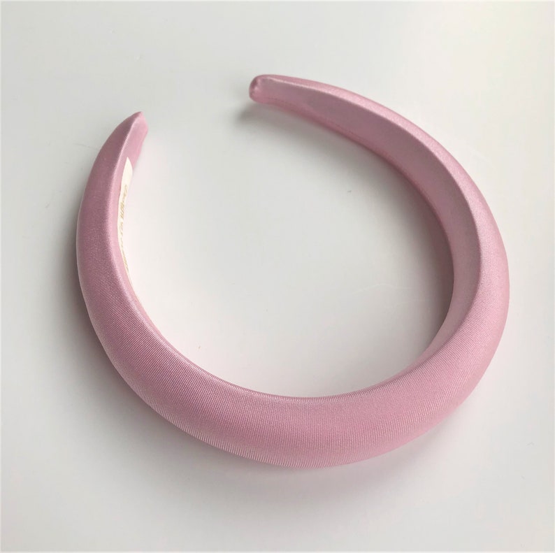 May include: A wide, pink satin headband. The headband is a simple, solid color and has a smooth, glossy finish.